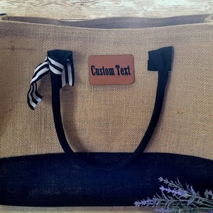 May include: A large, brown burlap tote bag with black trim and handles. The bag has a black and white ribbon tied to the handle and a brown leather tag with the text "Custom Text" on it.