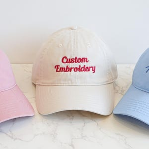 Puede incluir: Tres gorras de béisbol en colores pastel: rosa, crema y azul claro. Cada gorra tiene texto bordado en un color contrastante. La gorra rosa dice "By Millie", la gorra crema dice "Custom Embroidery" y la gorra azul dice "The Party".