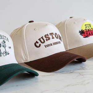 Puede incluir: Tres gorras de béisbol con diseños bordados. Una tiene "Margarita Social Club", otra "CUSTOM YOUR DESIGN" y la tercera "SUN SALT SAND". Cada gorra tiene una visera de color contrastante.
