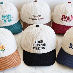 May include: A selection of corduroy baseball caps in different colours, each with embroidered text or designs. The caps have a cream-coloured crown and a contrasting brim, with text including "DAD" and "YOUR COUSTOM DESIGN".