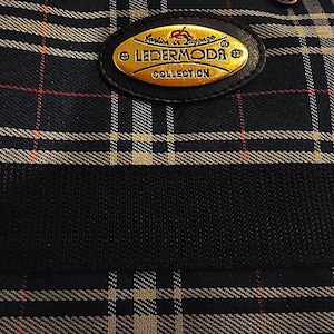 May include: A dark navy blue fabric bag featuring a classic plaid pattern with thin white and red lines. An oval-shaped gold-toned metal plaque is centred on the front, displaying the text LEDERMODA COLLECTION in black lettering.