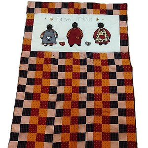 May include: A patchwork quilt in red, black, and gold squares. The top panel features three figures with the text "Forever Friends". Each figure is wearing an apron and holding a heart. The quilt is handmade.