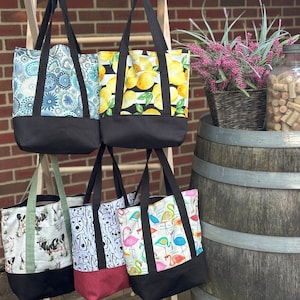 May include: Six different tote bags with various colorful patterns, including lemons, flamingos, and dogs, hanging on a wooden rack. The bags have black canvas bottoms and are displayed in front of a brick wall.