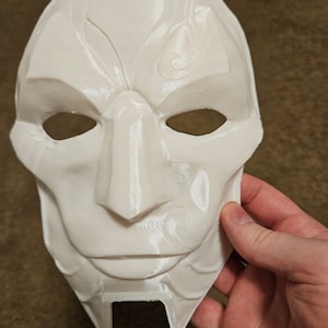 May include: White plastic mask with intricate details and a sharp, angular design. The mask has two eye holes and a small opening at the bottom of the chin.
