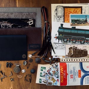 Könnte beinhalten: Eine Ansammlung von Vintage-Ephemera und Bastelmaterialien auf einer Holzoberfläche. Enthält Ledergeldbörsen, Stoffmuster, Metallverzierungen, Briefmarken und Zugillustrationen. Die Farbpalette ist erdig mit Braun-, Schwarz- und Cremetönen.
