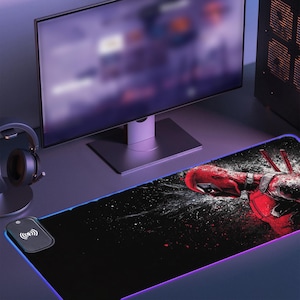 May include: Black rectangular gaming mouse pad with a red and black graphic of a superhero. The pad has a wireless charging area and a glowing blue and purple edge. A computer monitor and headphones are in the background.