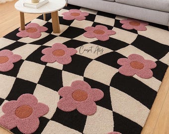 May include: A patterned rug featuring a black and cream chequered design with pink flower motifs. The rug is in a living room, with a grey sofa and a small round table. The text "Carpet Ally" is visible.