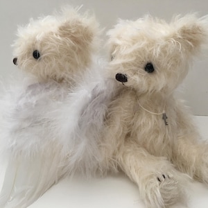May include: Two cream-colored teddy bears with black eyes and noses. One bear has a white feather wing attached. The bear on the right has a key pendant around its neck. The bears are sitting on a white surface.
