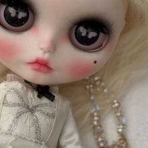 May include: A close-up of a doll's face with large, dark eyes reflecting a butterfly design. The doll has pale skin, rosy cheeks, and a small beauty mark. It wears a white dress with silver embroidery and a black cross necklace.