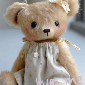 Puede incluir: Un oso de peluche beige que lleva un vestido blanco con estampado floral y una diadema de flores blancas. El oso tiene ojos marrones y una nariz marrón. El vestido tiene un botón blanco y el texto "hug me" está escrito en la parte inferior del vestido.