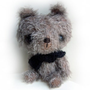 May include: A gray, furry teddy bear with a black knitted scarf. The bear is sitting on a white surface.