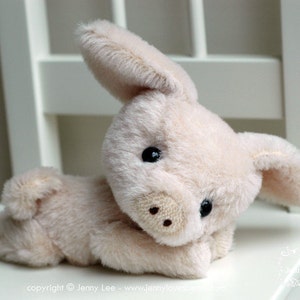 May include: A soft, plush, cream-colored pig toy with black eyes and a pink snout. The pig is lying on its side on a white surface.