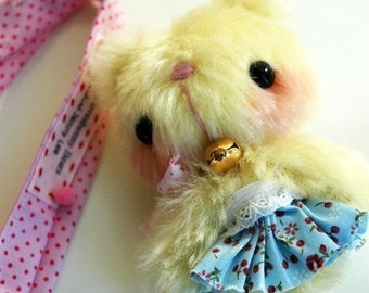 May include: A small, yellow plush toy bear wearing a blue and white floral dress with a white lace trim. The bear has a pink nose and black eyes and is wearing a gold bell around its neck.