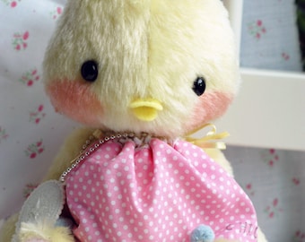 May include: A yellow plush toy chick wearing a pink polka dot dress with blue pom-poms. The chick has a yellow beak and blue and white pom-poms on its head.