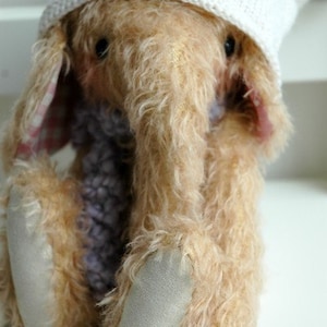 May include: A plush, light brown elephant toy wearing a white knitted hat with a pink and white gingham bow. The elephant has a long trunk and large, floppy ears.