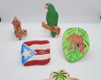 May include: A collection of ceramic souvenirs featuring a green parrot on a branch, a Puerto Rican flag, a frog on a leaf, a fire hydrant, and a sign that says "Puerto Rico". The items are painted in various colors.