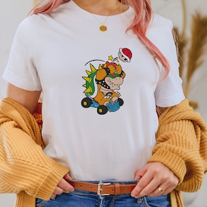 May include: A white t-shirt with a cartoon image of Bowser from the Super Mario franchise. Bowser is wearing a helmet and driving a kart. He is being hit by a shell.