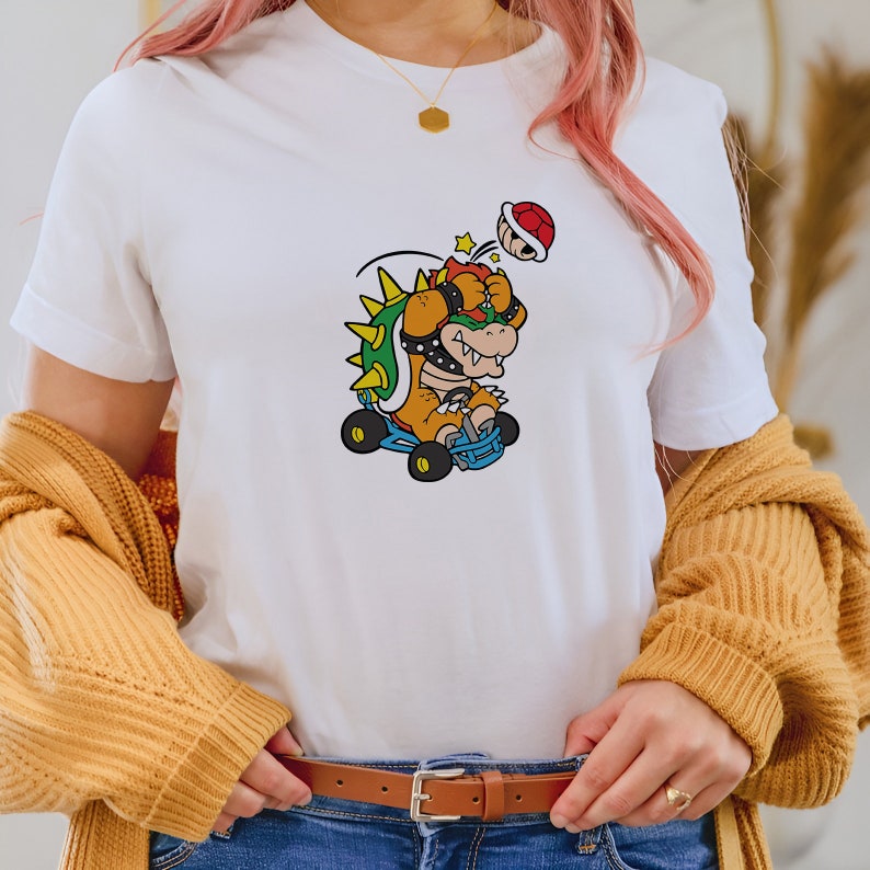 May include: A white t-shirt with a cartoon image of Bowser from the Super Mario franchise. Bowser is wearing a helmet and driving a kart. He is being hit by a shell.