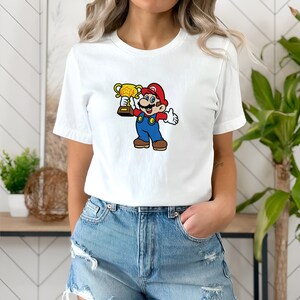 May include: White t-shirt with a cartoon image of Mario holding a gold trophy. Mario is wearing his signature red hat, blue overalls, and white gloves.