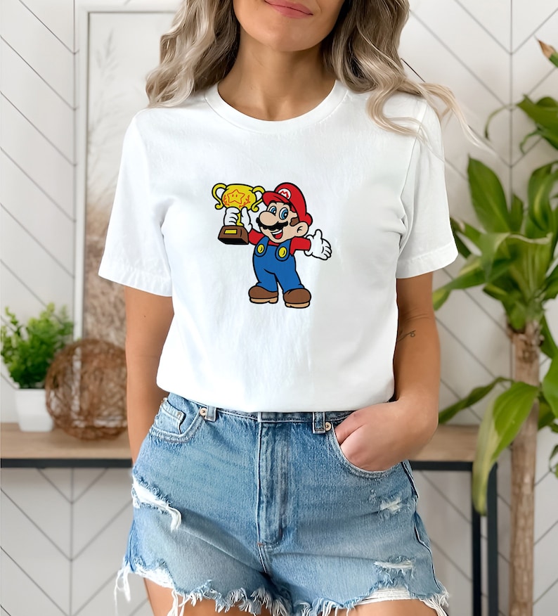 May include: White t-shirt with a cartoon image of Mario holding a gold trophy. Mario is wearing his signature red hat, blue overalls, and white gloves.