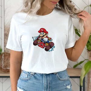 May include: White t-shirt with a graphic of Mario driving a kart. Mario is wearing a red cap and overalls and is holding a steering wheel. The kart is red and has a blue and white smoke trail behind it.