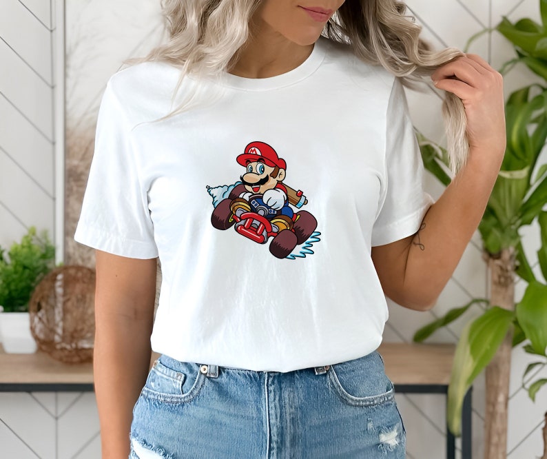 May include: White t-shirt with a graphic of Mario driving a kart. Mario is wearing a red cap and overalls and is holding a steering wheel. The kart is red and has a blue and white smoke trail behind it.