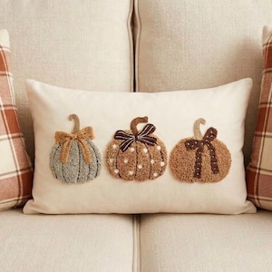 Peut inclure: Un coussin rectangulaire de couleur crème orné de trois citrouilles brodées. La citrouille de gauche est vert sauge avec un nœud beige, celle du centre est marron à pois blancs avec un nœud marron foncé, et celle de droite est beige avec un nœud marron.