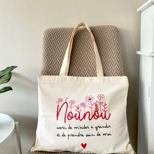 Può includere: Borsa tote color crema con testo ricamato rosso e design floreale. Il testo recita "Nounou" con le frasi "merci de m'aider à grandir et de prendre soin de moi" e un piccolo cuore rosso. La borsa ha manici color crema.