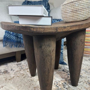 May include: A dark brown wooden stool with three legs. The stool has a rounded top and is sitting on a rug with a light brown and beige pattern. Two white books with the word "LONDON" printed on them are sitting on top of the stool.