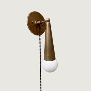 May include: A brass wall sconce with a cone-shaped shade and a white globe light bulb. The sconce has a cord with a black braided wire.