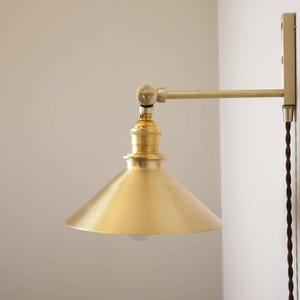 May include: A brass-toned wall-mounted lamp with a conical shade. The lamp features a pivoting arm and a decorative cord. The fixture is mounted on a rectangular plate against a white wall. The lamp's design is minimalist and industrial.