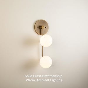 May include: A brass wall light with two spherical, glowing light bulbs. The fixture has a circular backplate and a vertical rod connecting the bulbs. Text on the image reads: Solid Brass Craftsmanship, Warm, Ambient Lighting.