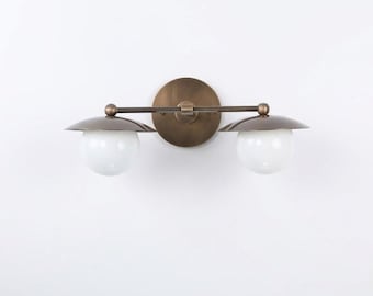 May include: A wall-mounted light fixture featuring a circular brass backplate and a horizontal bar supporting two downward-facing arms. Each arm holds a white spherical glass globe shade topped with a wide, flat, circular brass metal cover.
