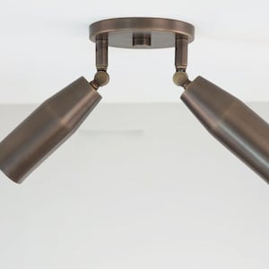 May include: Bronze-colored ceiling spotlight fixture with two adjustable heads. The fixture has a circular base and two cylindrical spotlights that can be angled. The spotlights are made of metal and have a smooth finish. This lighting fixture is suitable for indoor use.