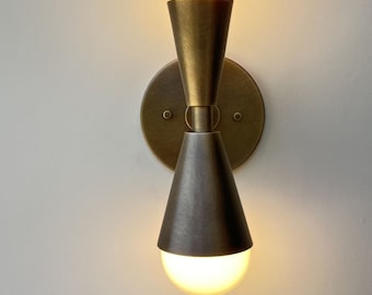 以下が含まれることがあります： 2つの電球が付いた真鍮製の壁掛け照明。照明は丸いベースと2つの円錐形のシェードを持っています。