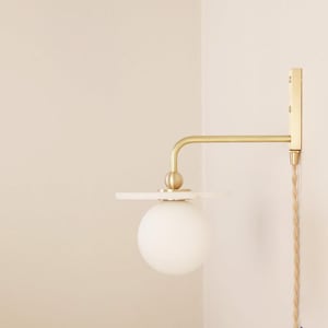 May include: A modern wall sconce with a brass finish, featuring a white globe light and a small, round shelf. The fixture is mounted on a light-colored wall, with a braided cord hanging down.