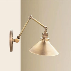 May include: A brass wall-mounted light fixture with a swivel arm and a cone-shaped shade. The light fixture has a vintage industrial style.