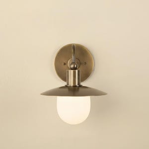 May include: A wall-mounted light fixture with a brass finish. The fixture features a circular backplate, a cylindrical body, and a downward-facing, saucer-shaped shade. A frosted, spherical bulb is visible.