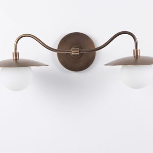 May include: A bronze-toned, two-light wall sconce. The fixture features a circular backplate, a curved arm, and two downward-facing, saucer-shaped shades. Each shade holds a spherical, white glass globe. The sconce is designed for indoor use.