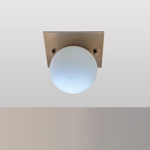 May include: A modern ceiling light fixture with a square brass base and a frosted glass globe.