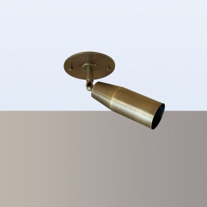 May include: A brass ceiling light fitting with a cylindrical shade that can be adjusted to point in different directions. The fitting has a round base with three screws.