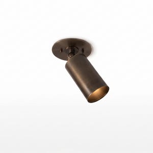 May include: A bronze-colored, adjustable spotlight fixture mounted on a white ceiling. The cylindrical light head is angled downward, with a warm glow emanating from the opening. The fixture is designed for directional lighting.