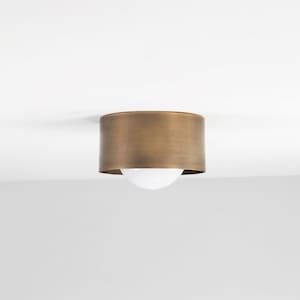 May include: A flush mount ceiling light fixture with a cylindrical, antique brass shade. The light has a white, spherical bulb. The fixture is mounted on a white ceiling.
