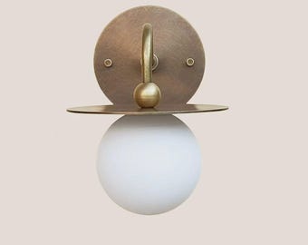 Op de afbeelding: Een moderne wandlamp met een ronde, messingkleurige achterplaat, een gebogen arm en een bolvormige, witte glazen lampenkap. De lamp heeft een klein, horizontaal plankje onder de achterplaat. Minimalistisch en elegant ontwerp.