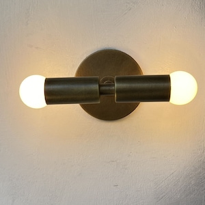 Op de afbeelding: Een messing wandlamp met twee gloeilampen. De lamp heeft een ronde basis en twee armen die naar de zijkanten uitsteken. De gloeilampen zijn aan en geven een warm wit licht.