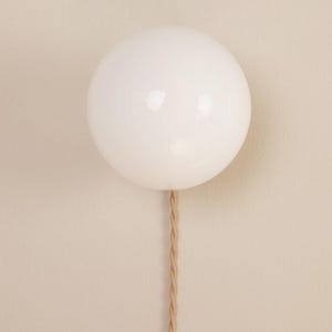 Op de afbeelding: Een witte, bolvormige lamp met een glanzende afwerking is op een beige muur gemonteerd. Een gedraaid, bruin koord loopt naar beneden. De lamp is een modern designelement.