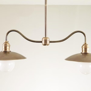 May include: A bronze-toned, two-light pendant lamp with a curved arm design. Each light features a brass-colored socket and a rounded, white glass bulb, with a matching bronze-colored shade. The lamp is suspended from a central rod.