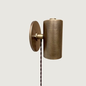 Op de afbeelding: Een messing wandlamp met een cilindrische kap en een ronde wandplaat. De lamp heeft een zwart snoer dat er vanaf hangt.