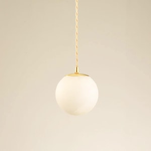 May include: A pendant light fixture featuring a white globe shade. The shade is suspended by a gold-colored metal fixture and a twisted beige cord. The light hangs against a neutral background.