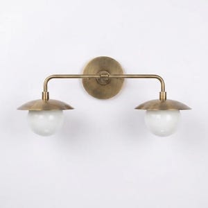 May include: A two-light wall sconce featuring a circular brass backplate, a horizontal brass arm, and two downward-facing brass shades. Each shade is paired with a spherical white glass globe bulb, creating a balanced and symmetrical lighting fixture.